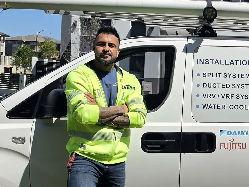 Davad Cala, owner of Union Air Conditioning, in a hi-vis jacket with the Union logo, standing with arms crossed beside the Union van