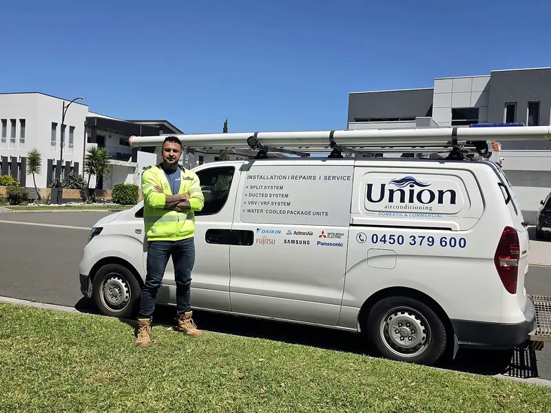 Union Air Conditioning technician in a hi-vis jacket with the Union logo, standing beside the white Union van with its roof ladders on a Sydney street