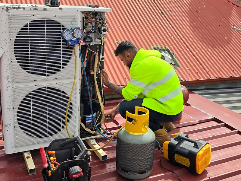 Union Air Conditioning technician in a hi-vis jacket crouching beside an opened outdoor unit on a roof, checking the wiring with a gauge set connected