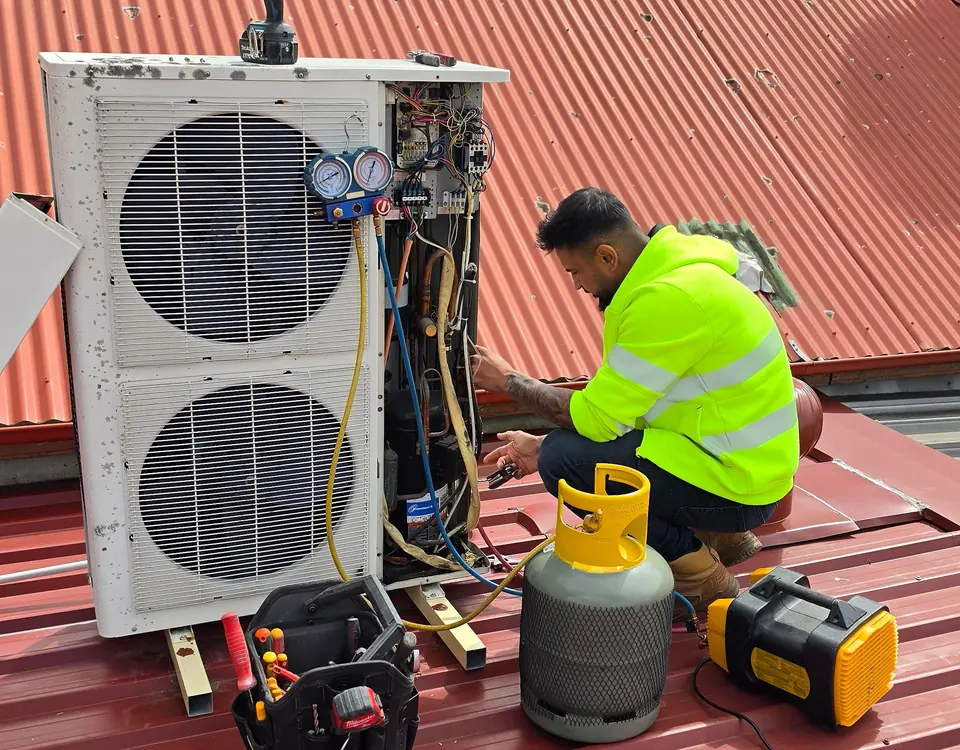 Union Air Conditioning technician in a hi-vis jacket repairing a large outdoor air conditioning unit on a corrugated iron roof, with a gauge set, refrigerant cylinder and recovery unit beside him